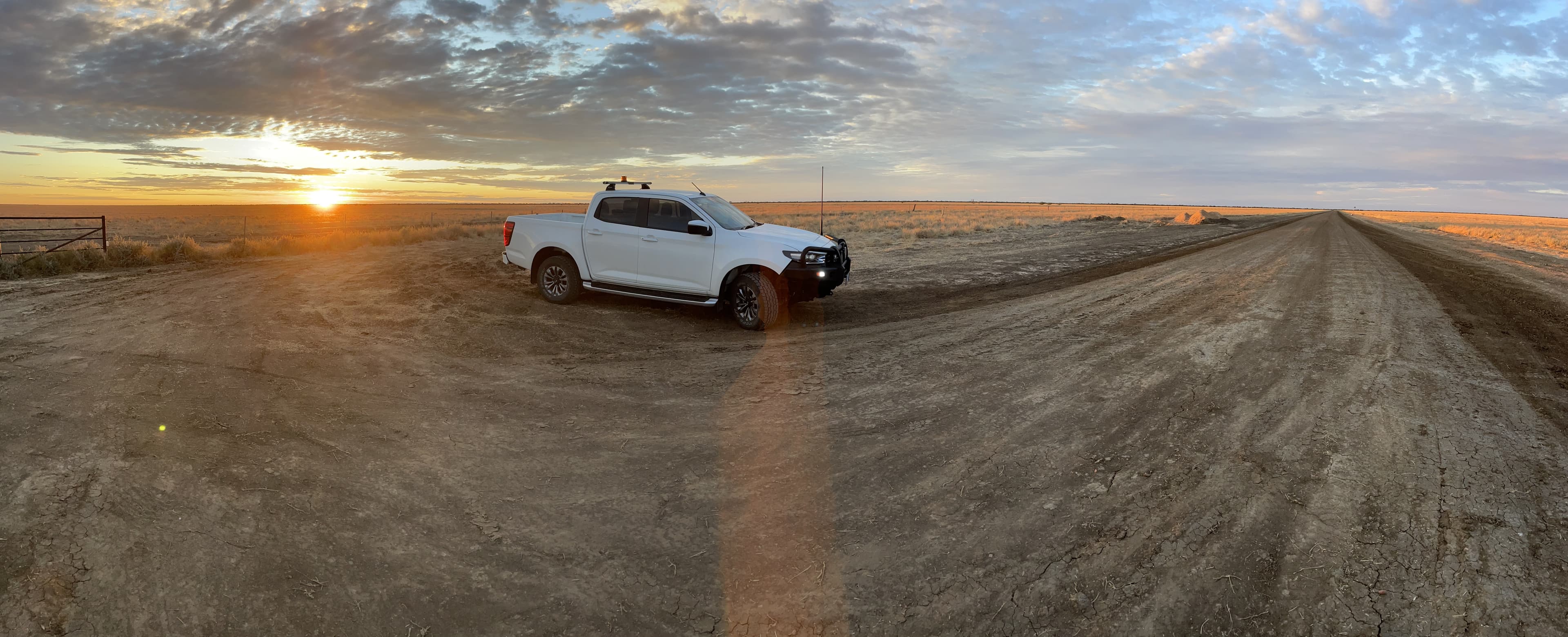 A white utility vehicle with a roof beacon parked beside a long dirt road on open plains at sunrise