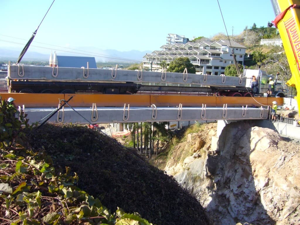A crane lowers a precast deck unit across the rock cutting, with the next unit waiting on a truck