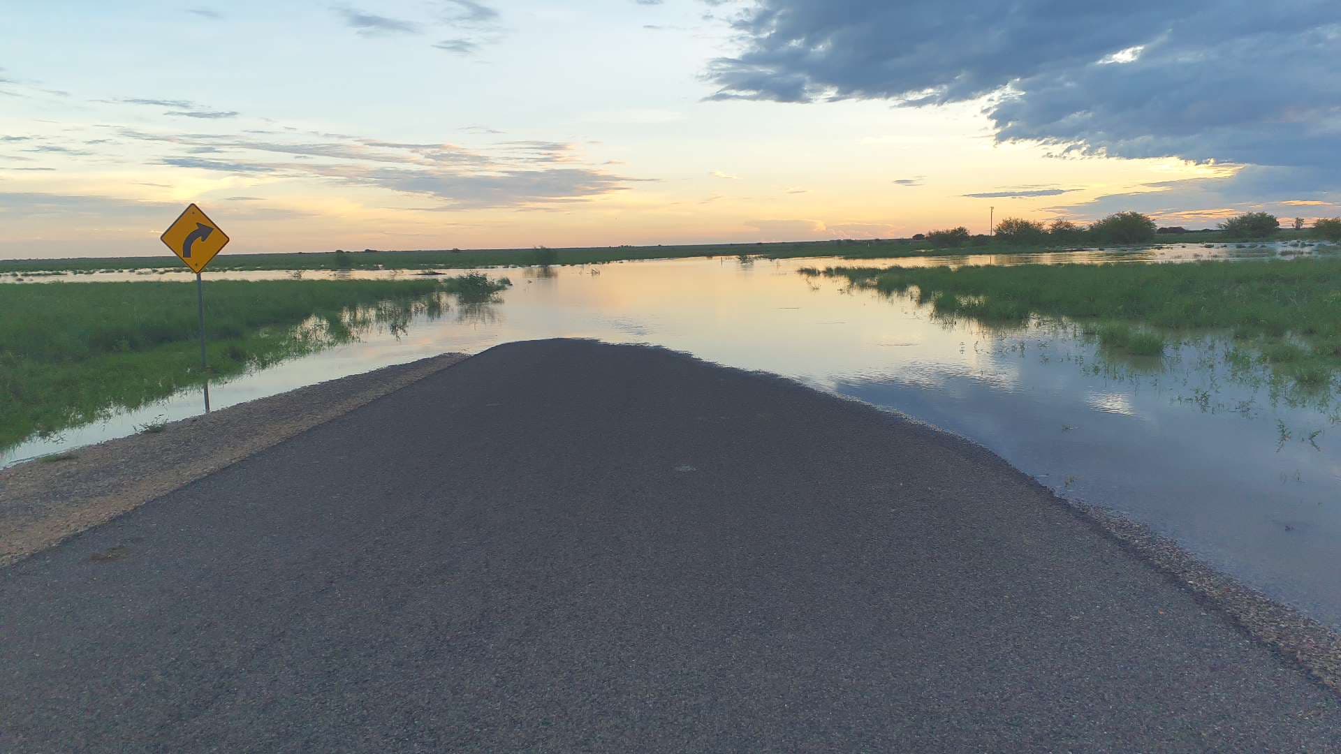 Floodwater over the edge of a sealed road at dusk, beside a curve warning sign