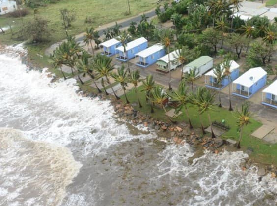 Aerial view of waves washing over the Bowen foreshore beside a row of cabins and palm trees