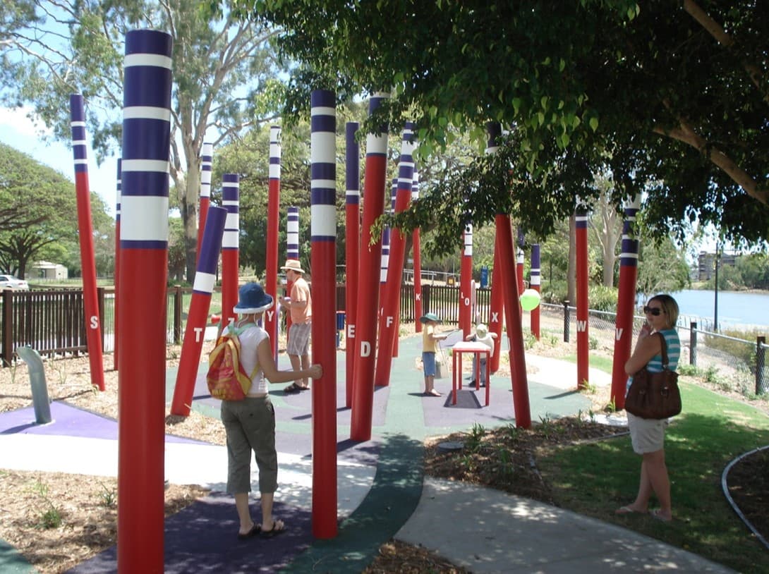 Visitors among the tall painted poles of the All Abilities Playground beside the river at Riverway