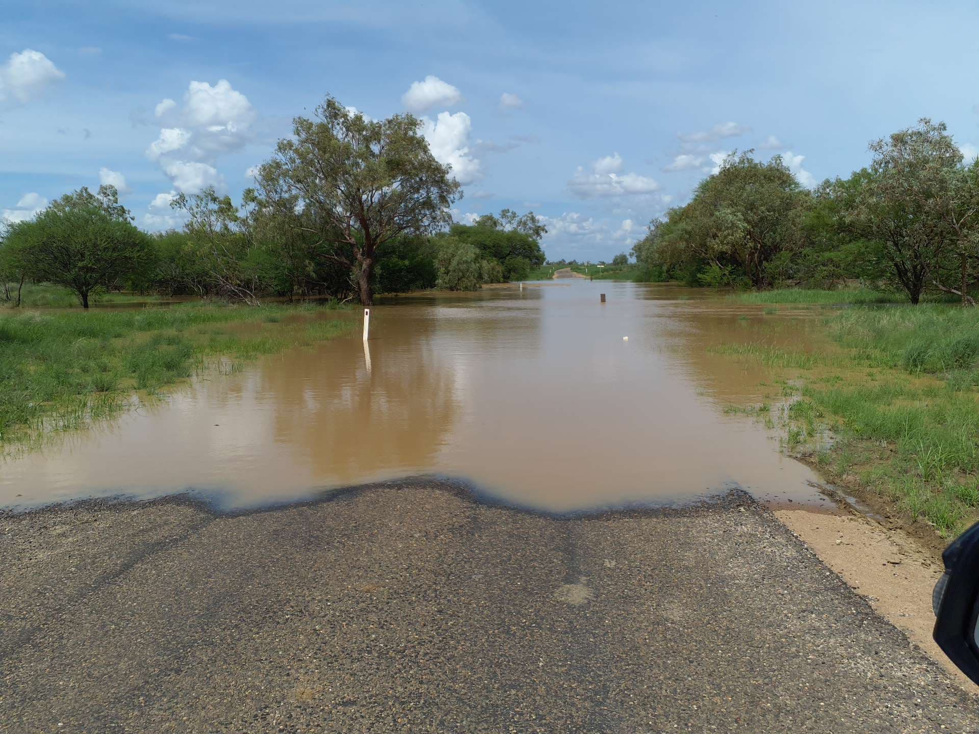 Floodwater flowing over a sealed outback road, with a guide post marking the road edge